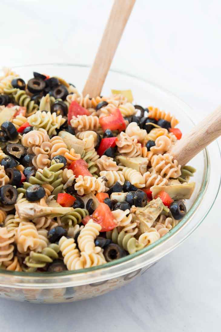 Una fotografía de cerca lateral de una receta de ensalada de pasta fácil con dos espátulas de madera en un tazón de vidrio.