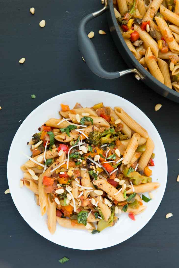 Overhead of pasta with vegetables served on a white plate with pine nuts.