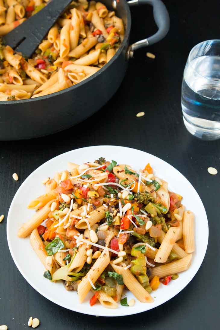 A side photograph of pasta with vegetables and scattered pine nuts. The main serving pot is in the background.