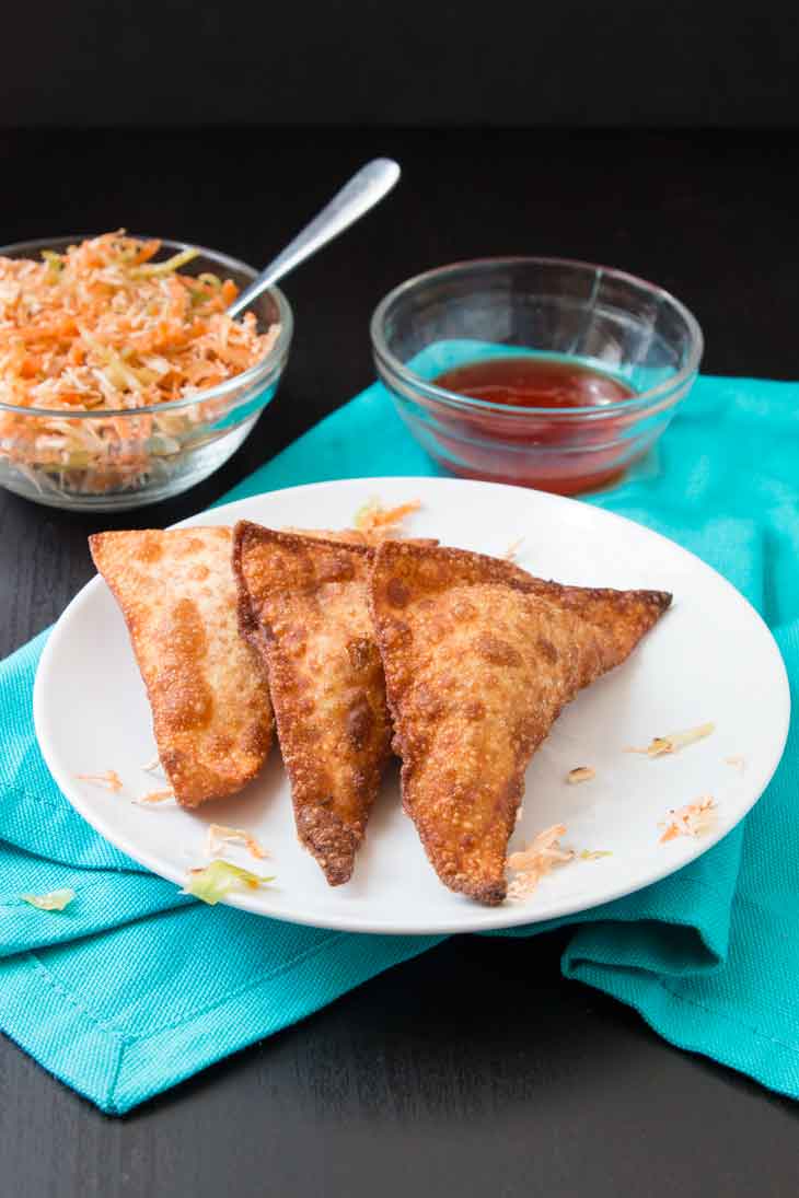 A side photograph of three vegan crispy wontons sitting on a round white plate and blue napkin, with dipping sauce and filling in the background. 