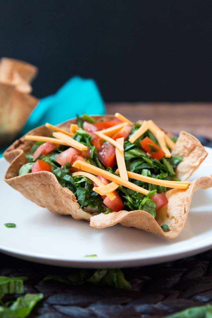 A photograph of a layered taco salad using a baked tortilla bowl. It's filled with black beans, spinach, tomatoes, and cheese.