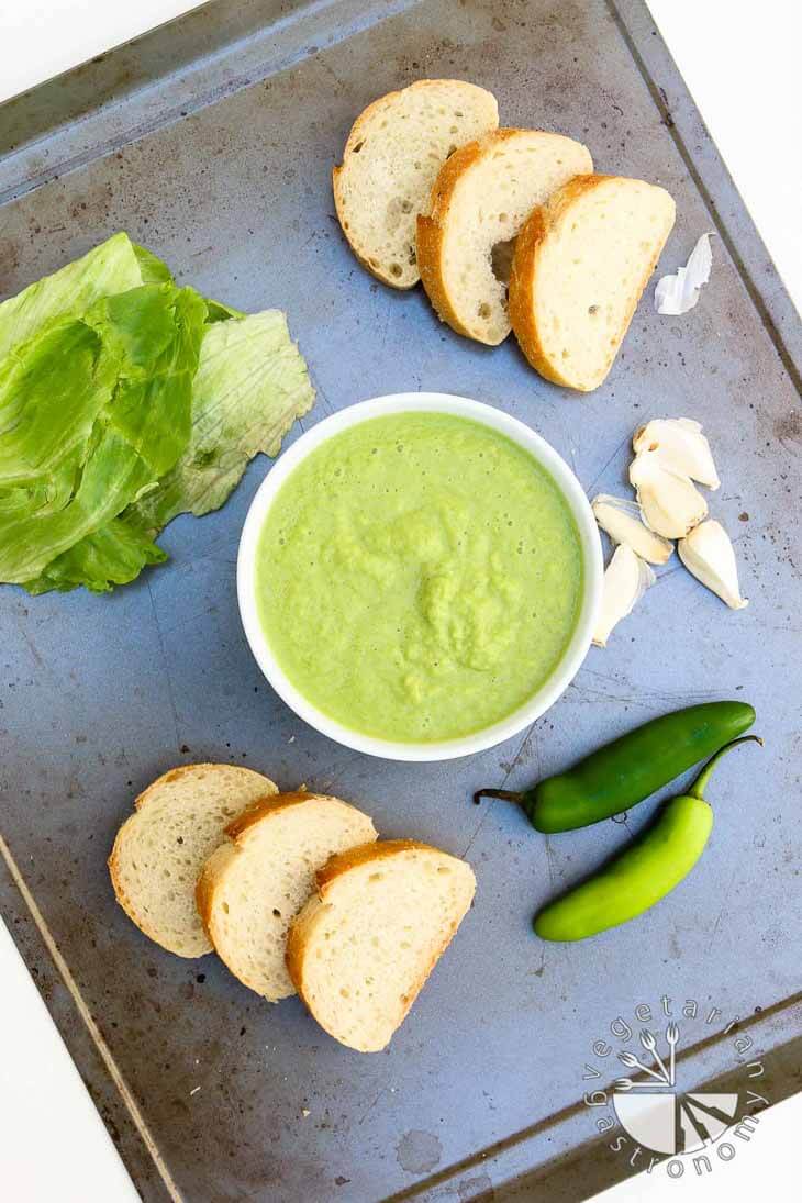 An overhead view of a baking sheet pan with a bowl of peruvian green sauce in the center, slices of french bread, garlic cloves, jalapenos, and lettuce.