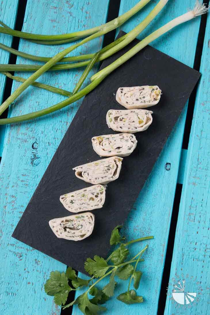 An overhead photograph of a black rectangular tray with vegan mexican tortilla pinwheels. There are green onions and cilantro off to the side sitting on a turquoise wooden palate.