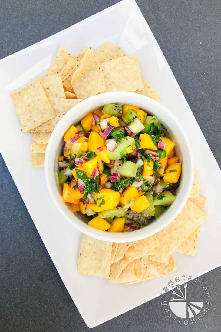 An overhead photograph of a white bowl filled with fresh mango kiwi salsa. It's sitting on a rectangular plate with chips around the side.