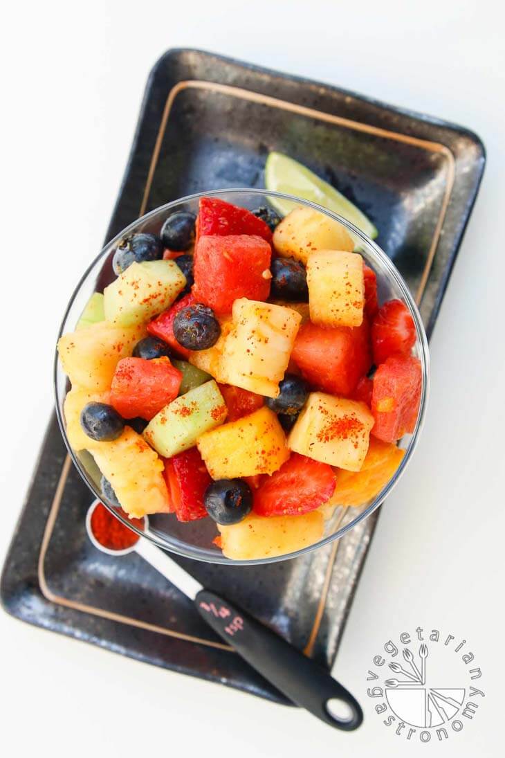 An overhead view of spicy fruit salad in a clear glass bowl. The bowl is sitting on a black rectangular plate with some lime and chili powder.