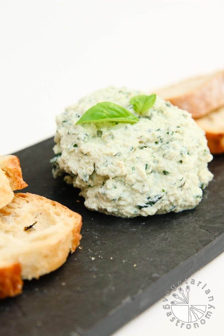 A side view of a small cheese plate consisting of garlic basil vegan ricotta cheese with slices of french bread. It's sitting on a black tray.