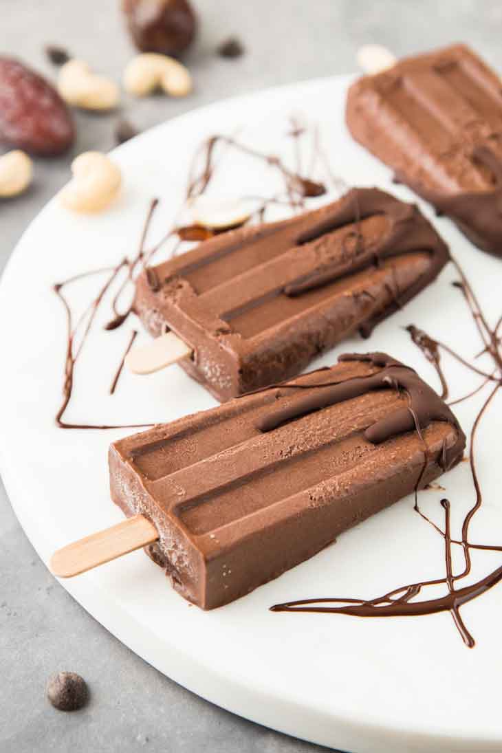 A side photograph of fudgesicles with chocolate drizzled on top. There are cashews and dates in the background.