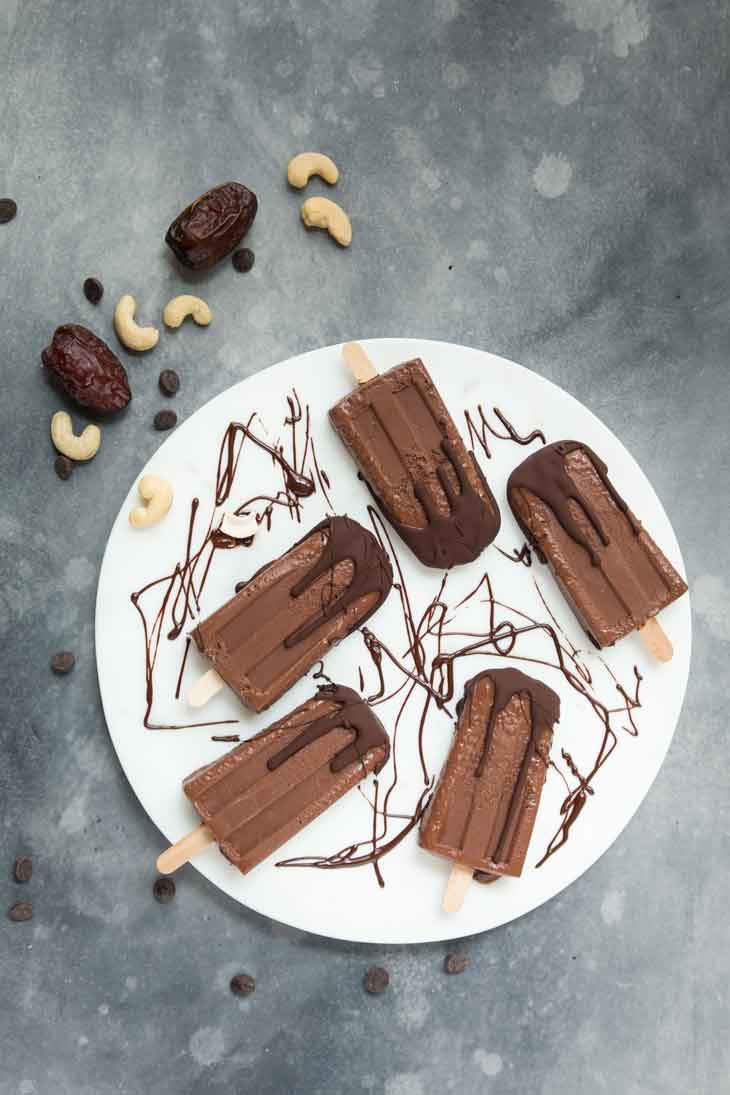 Overhead photograph of fudgesicles sitting on a white marble round board. There are dates, cashews, and chocolate chips scattered around.