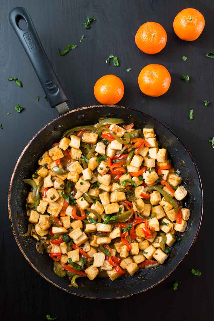 Overhead photograph of orange tofu stir-fry served on a frying pan with three navel oranges off to the side.