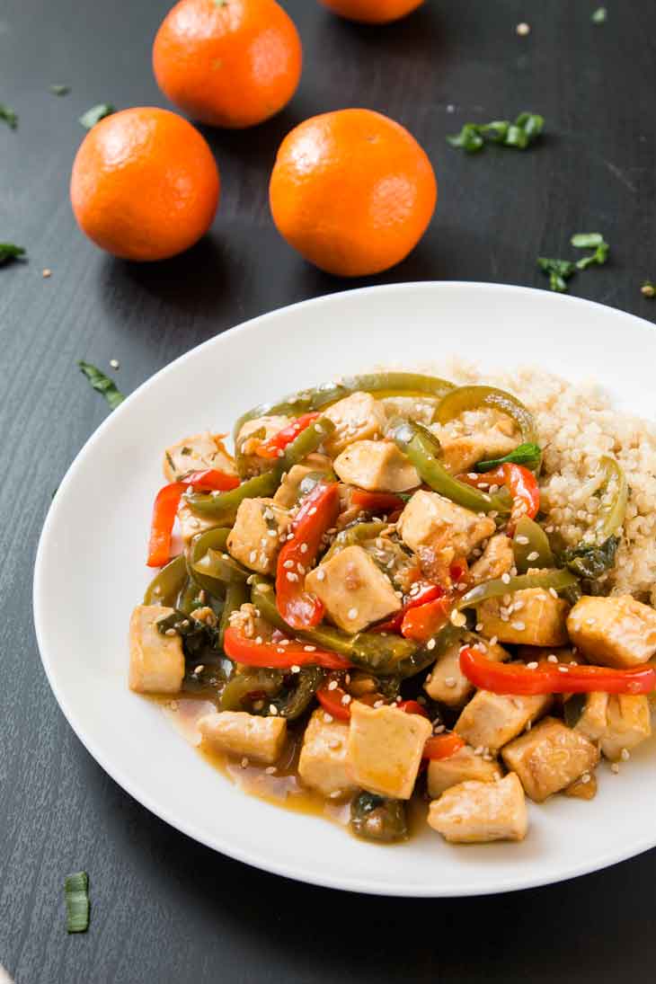 A side photograph of orange tofu stir-fry served over quinoa on a white plate. There are three navel oranges in the background.