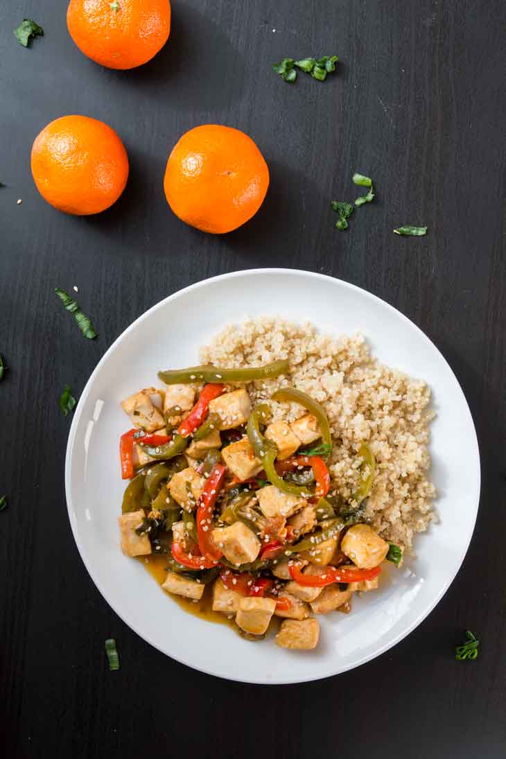 An overhead photograph of orange tofu stir-fry served with cooked quinoa on a white round plate. There are three navel oranges off to the side.