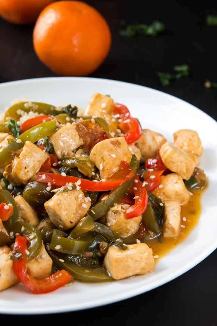 A close-up photograph of orange tofu stir-fry with peppers served on a white round plate. There are navel oranges in the background.