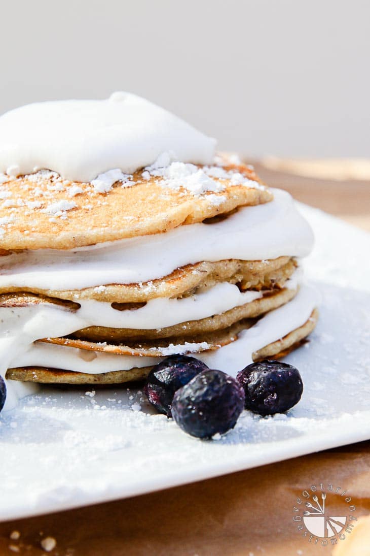 A side view photograph of four stacked banana macadamia nut pancakes with coconut whip cream. 