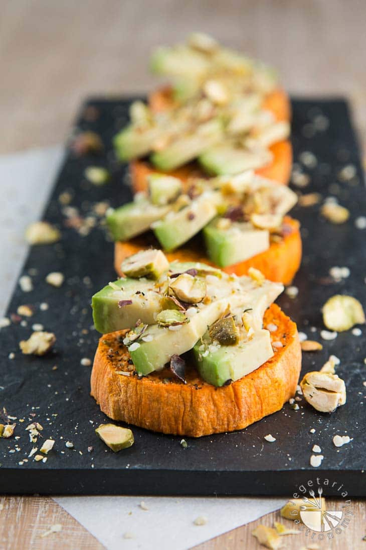 A front view of four sweet potato avocado toasts topped with hemp seeds and roasted pistachios. The toasts are sitting on top of a rectangular black board.