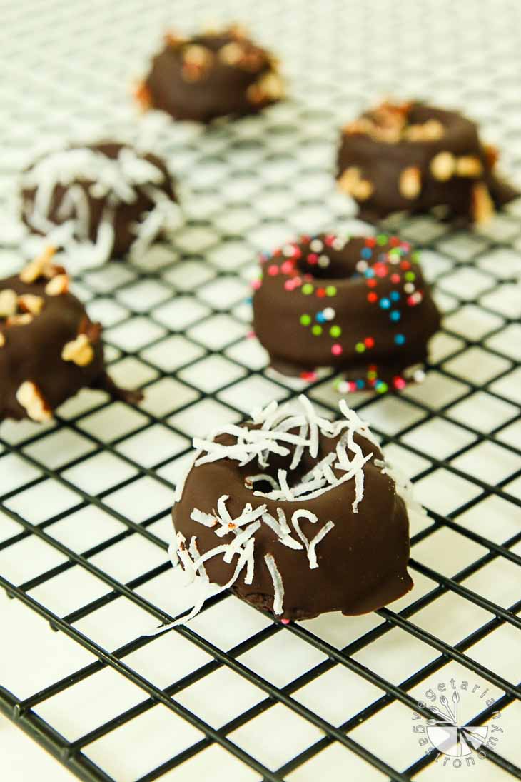 A 45 degree angle of vegan brunch recipes consisting of mini chocolate gluten-free donuts sprinkled with coconut, pecans, and sprinkles. They are sitting in a single layer on a cooling rack.
