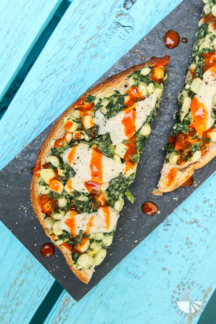 An overhead photograph of creamy corn and spinach toast with hot sauce drizzled on top. The toast is sitting on a rectangular black trayon a turquoise wooden palate. 