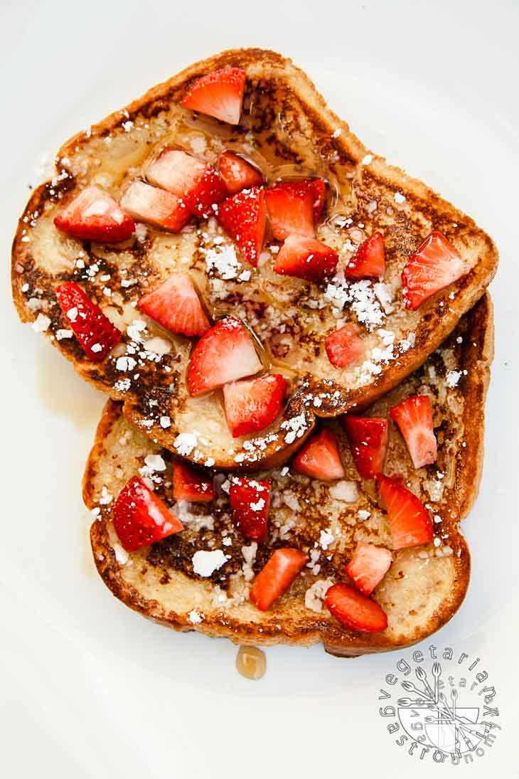 An overhead photograph of french toast. there are two toasts partially on top of each other, topped with maple syrup, powdered sugar, and diced strawberries. 