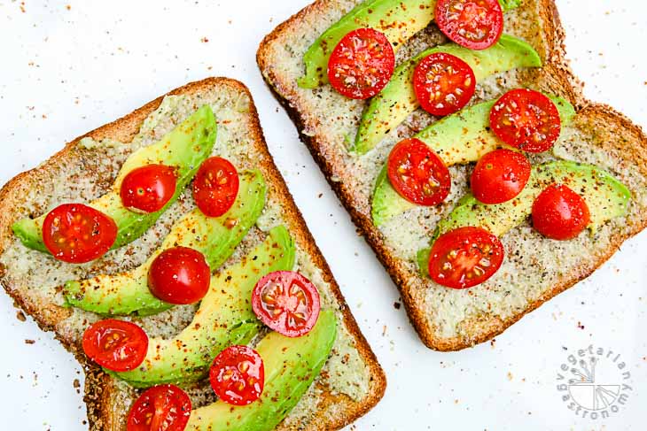 Overhead photograph of two avocado cherry tomato toasts sitting on a white background and topped with black pepper and salt. 