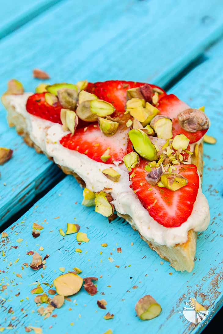 Cinnamon-infused mascarpone on focaccia topped with strawberries and roasted pistachios, on a turquoise wooden palate.