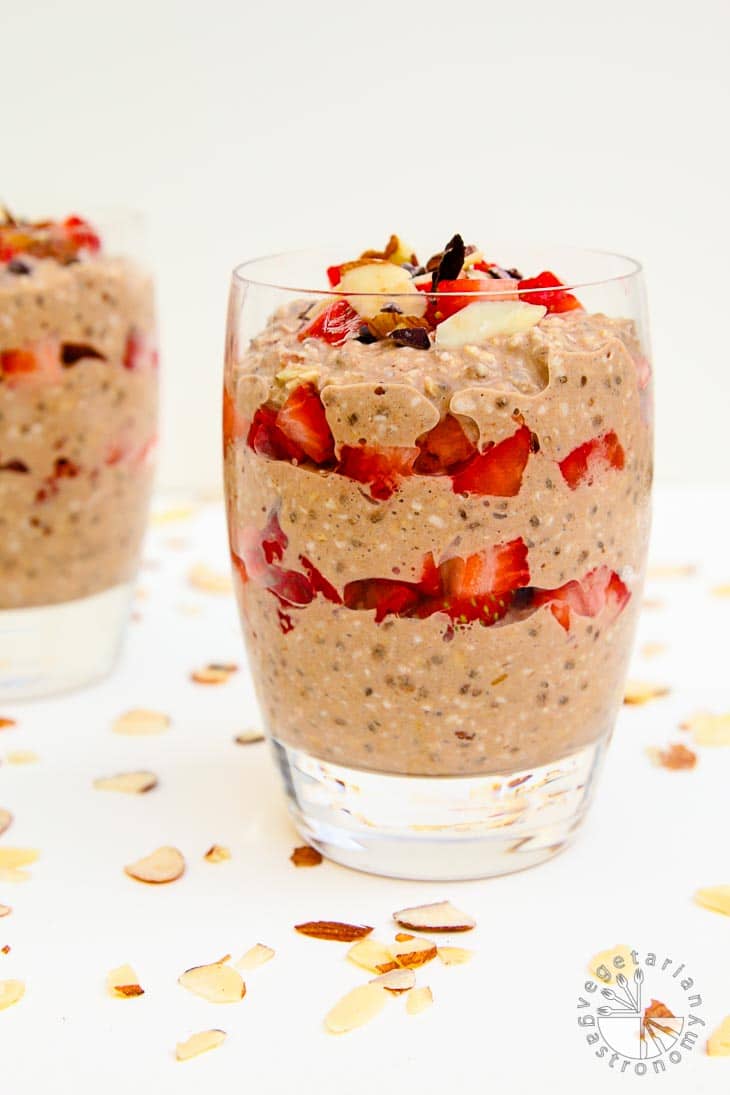 A side view photograph of two cups of chocolate strawberry almond overnight oats. The cups are sitting on a white board with almond slivers as garnish.