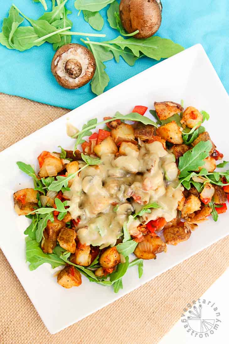 Overhead photograph consisting of of roasted potatoes, peppers, onions, and arugula, topped with a mushroom gravy on a white rectangular plate. There is a turquoise napkin, raw mushrooms, and arugula off to the side for garnish.