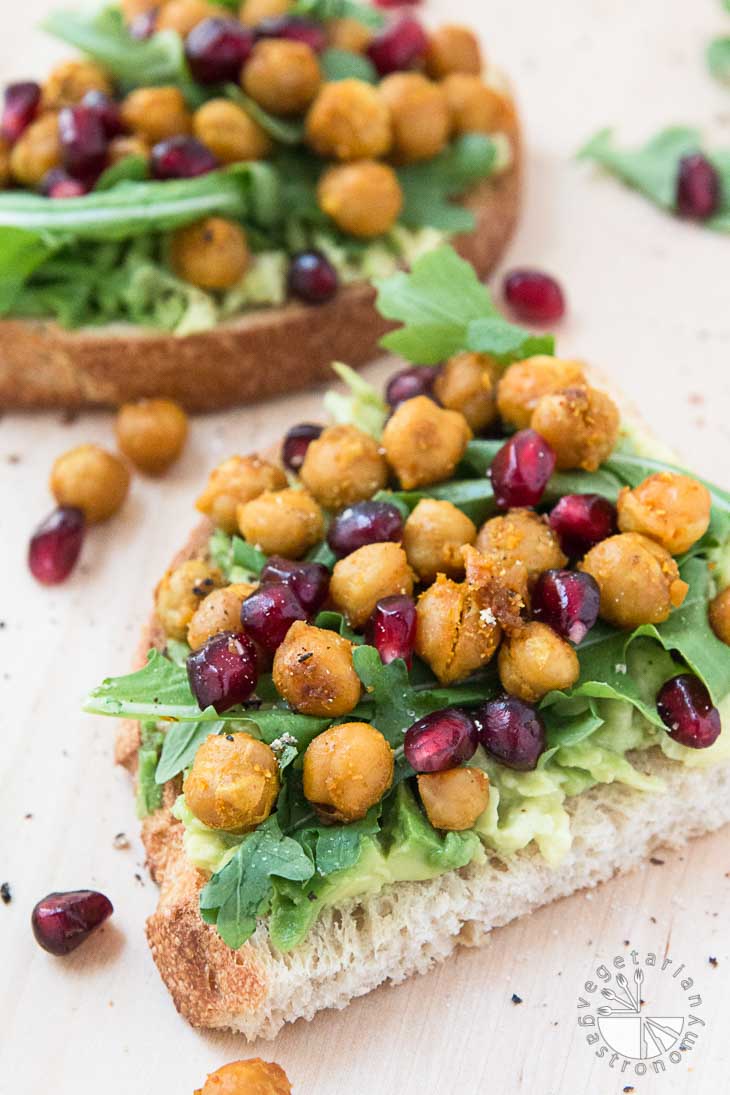 A 45 degree angle photograph of two loaded toast layered with avocado, arugula, roasted chickpeas, and pomegranate seeds. 