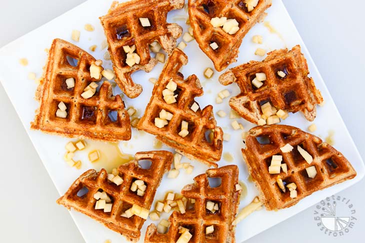 Overhead shot of apple cinnamon waffles cut into quarters and placed on a square white plate. The waffles are topped with maple syrup and diced apples.