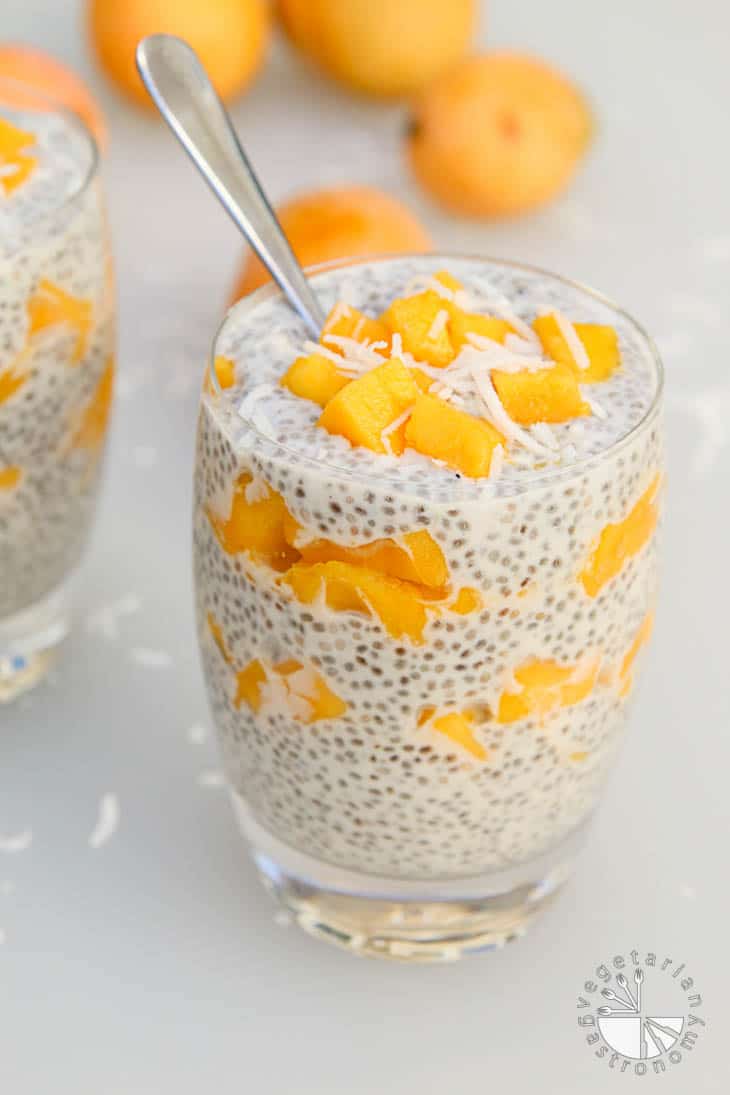 A 45 degree photograph of a glass cup layered with coconut chia pudding and freshly diced mango, topped with fresh coconut. The vegan brunch recipes have a spoon sitting in the cup.