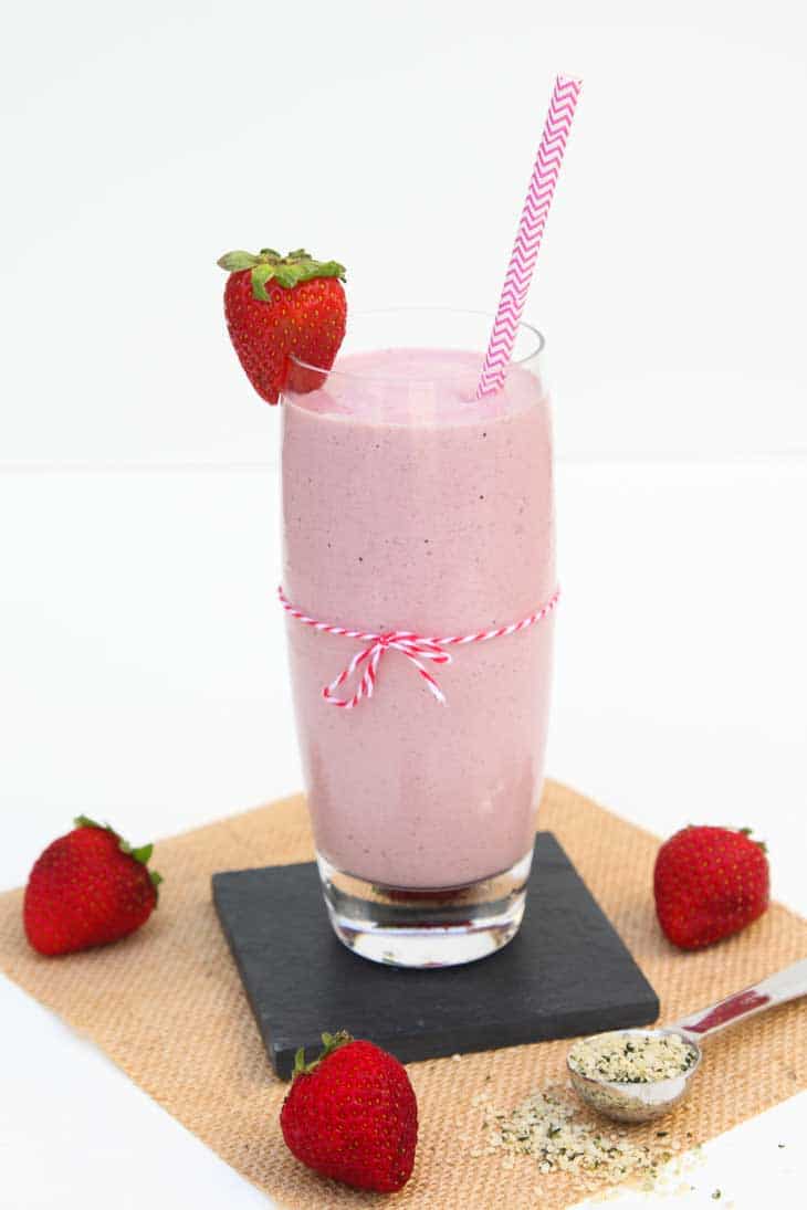 Side photograph of a tall glass of Strawberry Milkshake recipe. There's a pink straw, strawberries, and hemp seeds.