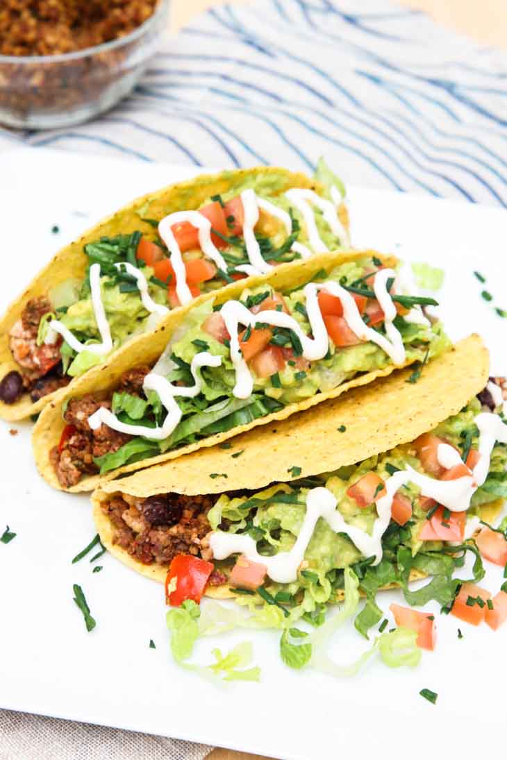 A 45 degree photograph of three loaded black bean tofu tacos stacked side-by-side on a white square plate.