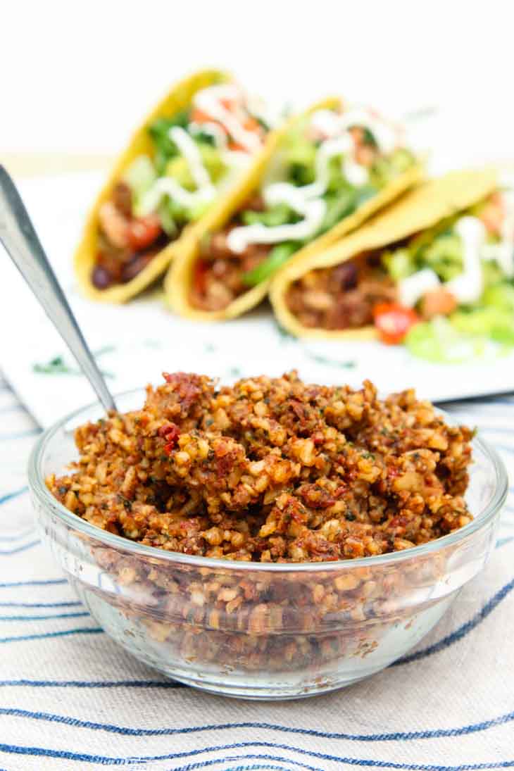 "Meatless" sun-dried walnut vegan crumble in a glass bowl with loaded tofu tacos assembled in the background.