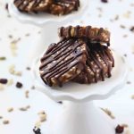 Portrait size image of No-Bake Chewy Chocolate Date Cookies. They are made with dates, seeds, and dried fruit, with chocolate drizzled on top. The date cookies are allergen-friendly and are gluten-free, dairy-free, soy-free, eggless, and vegan. They are sitting on a white cupcake stand with the seeds and dried fruit mixture sprinkled around it.