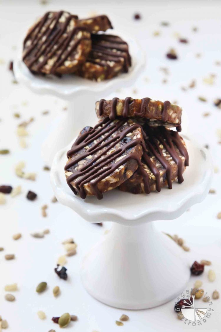 Portrait size image of No-Bake Chewy Chocolate Date Cookies. They are made with dates, seeds, and dried fruit, with chocolate drizzled on top. The date cookies are allergen-friendly and are gluten-free, dairy-free, soy-free, eggless, and vegan. They are sitting on a white cupcake stand with the seeds and dried fruit mixture sprinkled around it. 