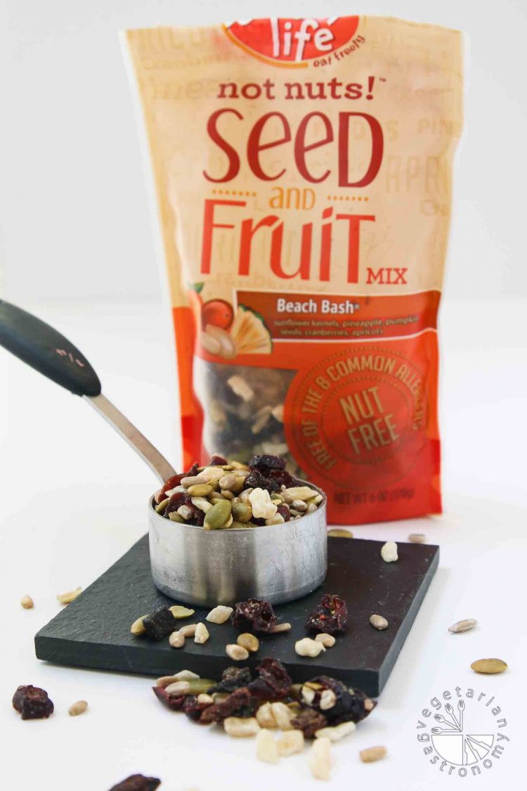 This picture of Enjoy Life Beach Bash seed and dried fruit mixture. the seed and dried fruit mixture used in my No-Bake Chewy Chocolate Date Cookies. The mix is sitting in a measuring cup with the bag behind it. It's soy-free, nut-free, gluten-free, and vegan.