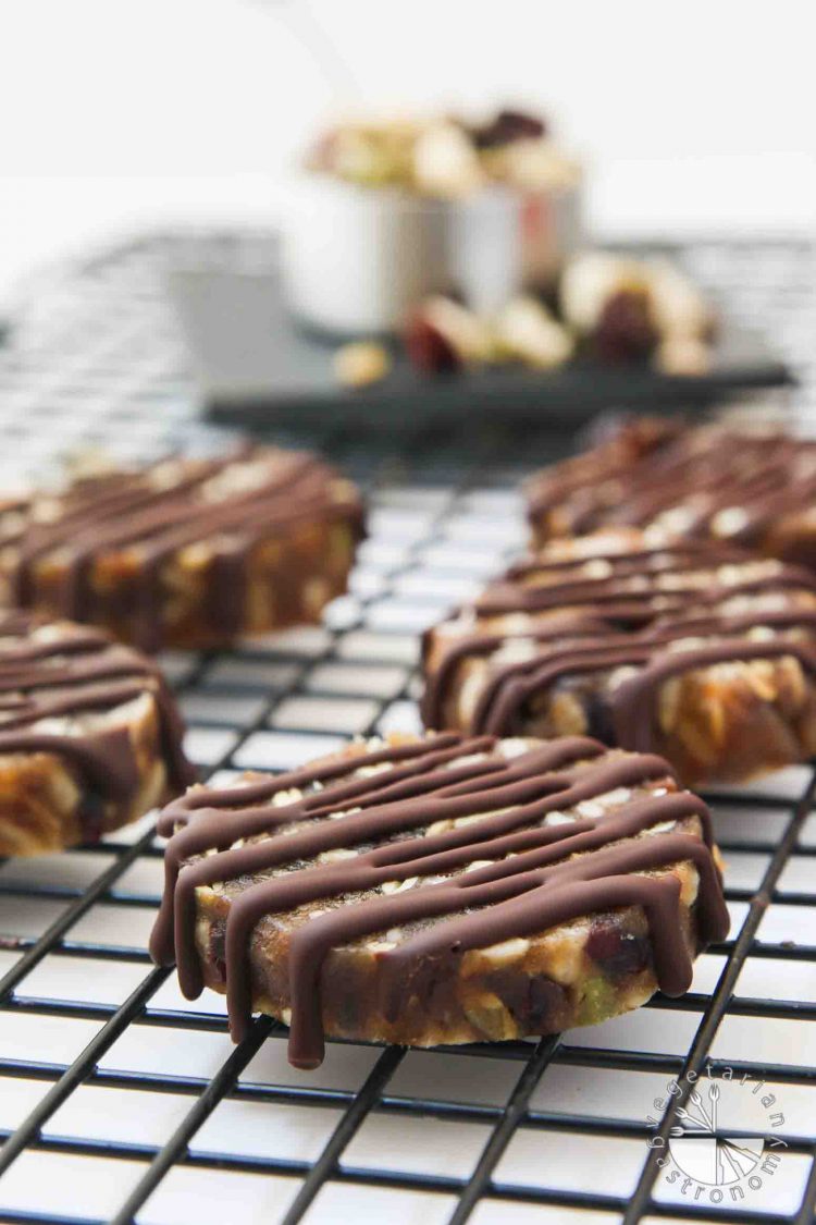 Portrait picture of No-Bake Chewy Chocolate Date Cookies sitting on a black cooling rack, taken at about 45 degrees. The seed and dried fruit mixture used for the recipe along with the mix bag sitting behind it blurred in the background. These healthy cookies are vegan, gluten-free, and nut-free.