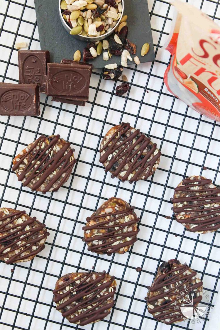 This image is an overhead shot of my healthy No-Bake Chewy Chocolate Date Cookies, Enjoy Life brand Boom Choco Boom Dark Chocolate Bars, and their Beach Bash Seed and Dried Fruit Mix. The easy cookies are vegan and gluten-free and are sitting on a black cooling rack. 