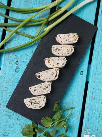 An Overview Shot of Six Vegan Mexican Tortilla Pinwheels Made with Cream Cheese. The Tortilla Pinwheels are Sitting on Top of a Black Rectangular Plate with some Green Onions and Cilantro off to the side for Garnish. The Backdrop is a Wooden Turquoise Palate.