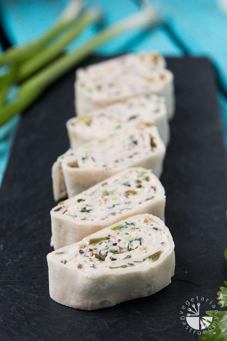 A 45 Degree Shot of Six Vegan Mexican Tortilla Pinwheels. They are sitting on a Black Plate with some Cilantro Blurred in the Background for Garnish. 