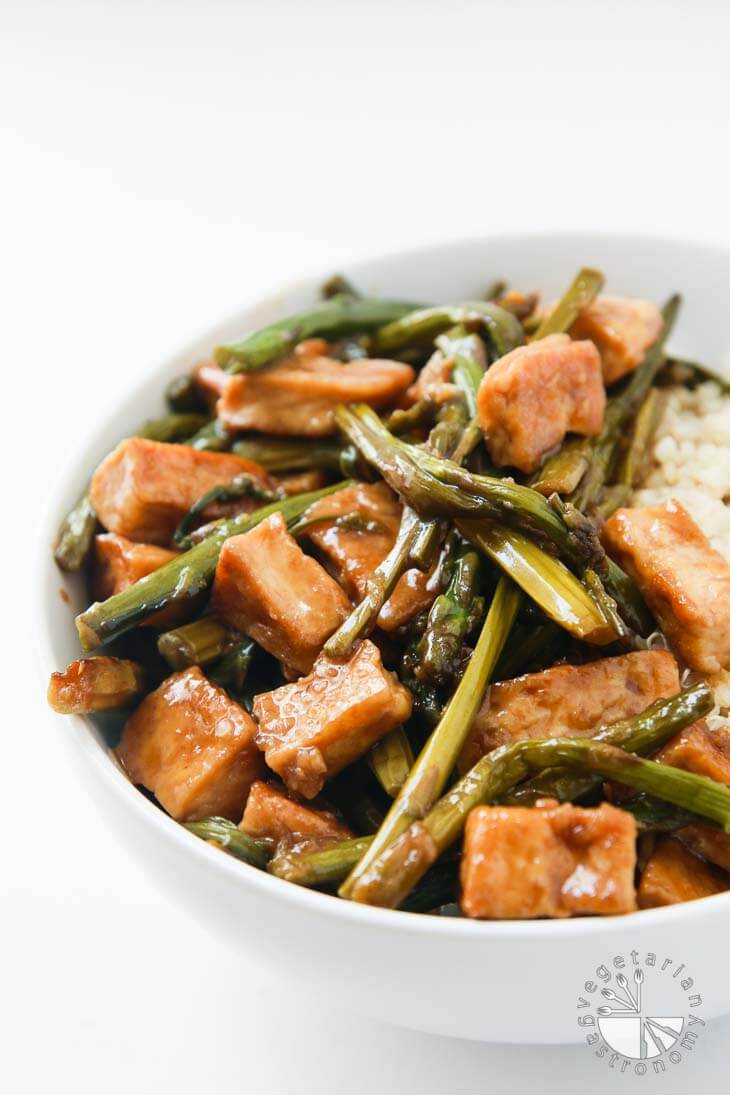 A closeup front photograph of a partial white bowl filled with Teriyaki tofu and asparagus over cooked quinoa. The bowl is sitting on a white board. 