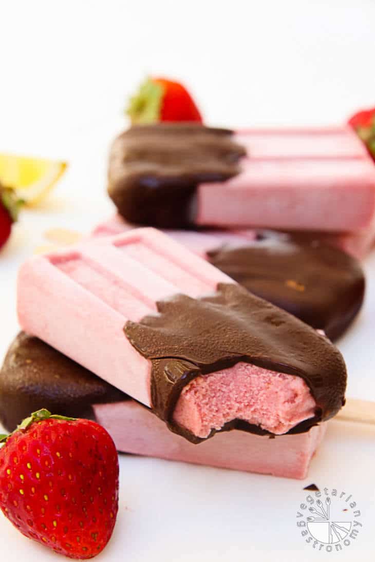 A side view of dark chocolate covered strawberry cream popsicles. There are four popsicles layered on top of each other with a bite taken out of it and strawberries as garnish.