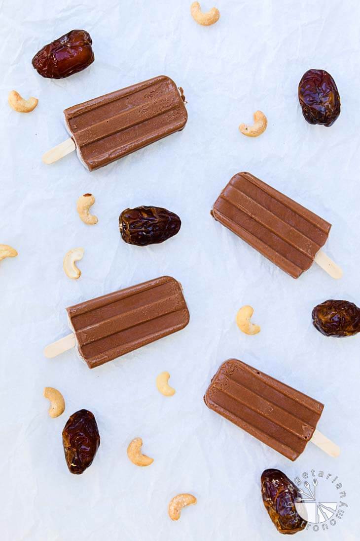 Overhead photograph of four vegan chocolate fudge popsicles with dates and cashews garnished around the sides.
