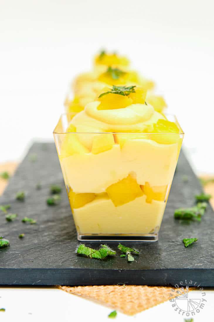 A front photograph of mango mouse parfait in square glass cups sitting on a black palate. There's mint around the sides as garnish.