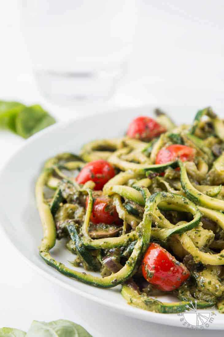 A side photograph of spiralized pesto zucchini spaghetti with tomatoes and mushrooms on a white plate.