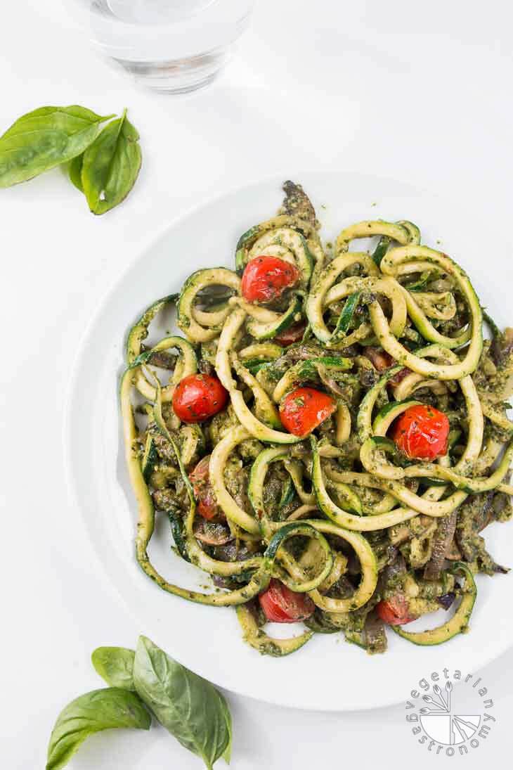 Overhead photograph of pesto zucchini spaghetti recipe on a white plate with a glass of water and fresh basil leaves off to the side.