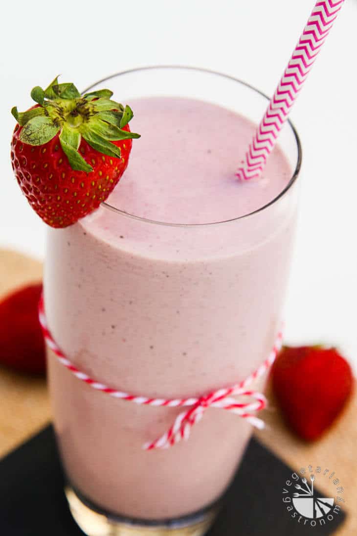 A tall glass of strawberry hemp milkshake with a pink and white straw. The glass has a string tied around it and a strawberry hanging on the lip.