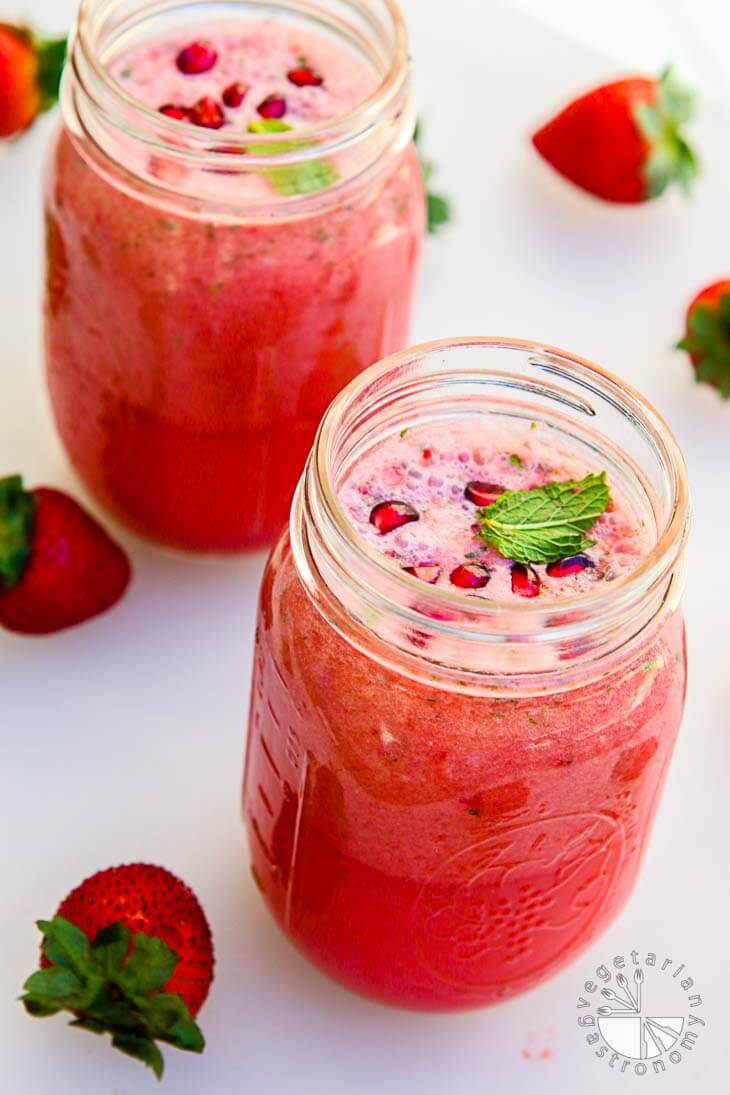 There are two mason jars filled with pomegranate strawberry lemonade with pomegranate pearls and some mint leaves on top. There are strawberries around the sides of the jars for garnish.