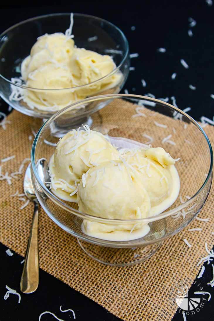 A side photograph of two glass cups filled with tropical creamy soft serve. They are sitting on a mesh paper with shredded coconut and spoon for garnish.