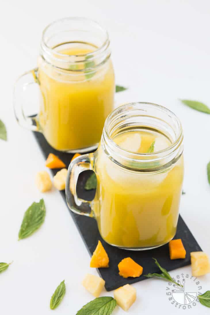 A 45 degree photograph of two mason jars filled with pineapple mango agua fresca recipe. There are pieces of fresh fruit and mint leaves garnished around the sides.