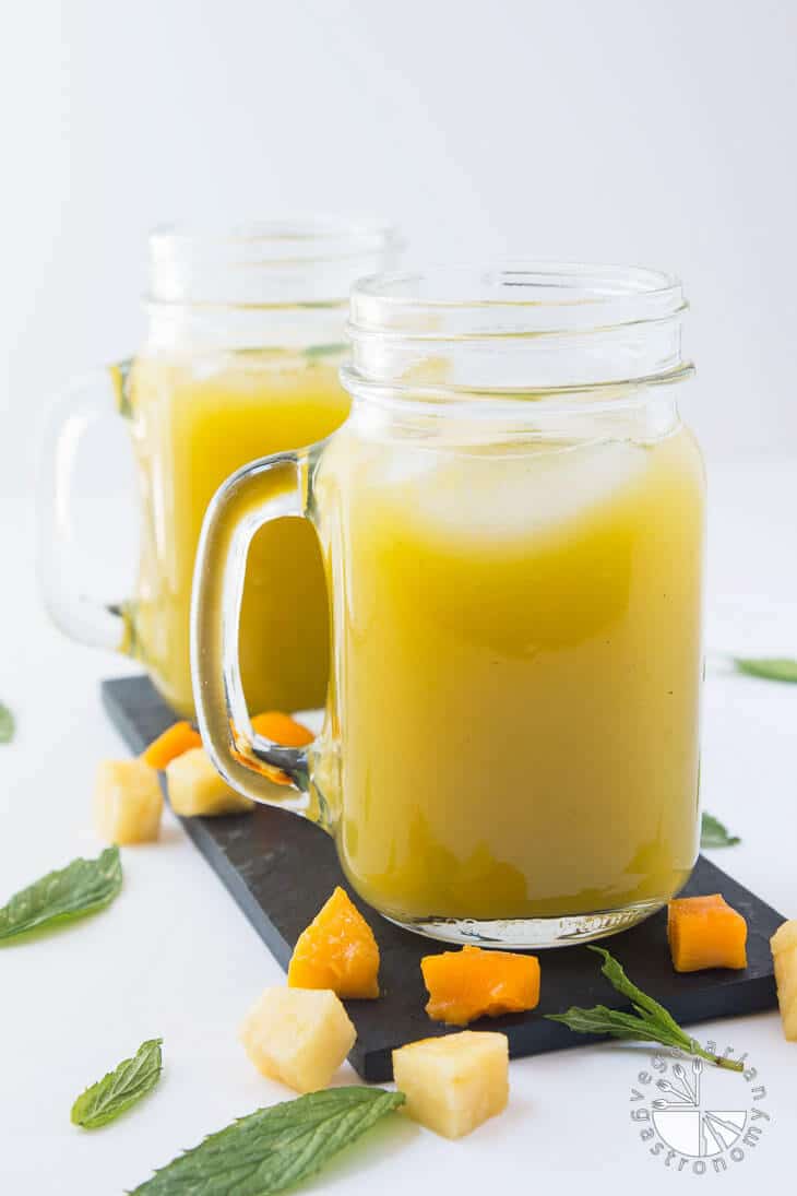 A side photograph of two mason jars filled with pineapple mango agua fresca recipe. There are pieces of mint, mango, and mint leaves garnished around the jars.