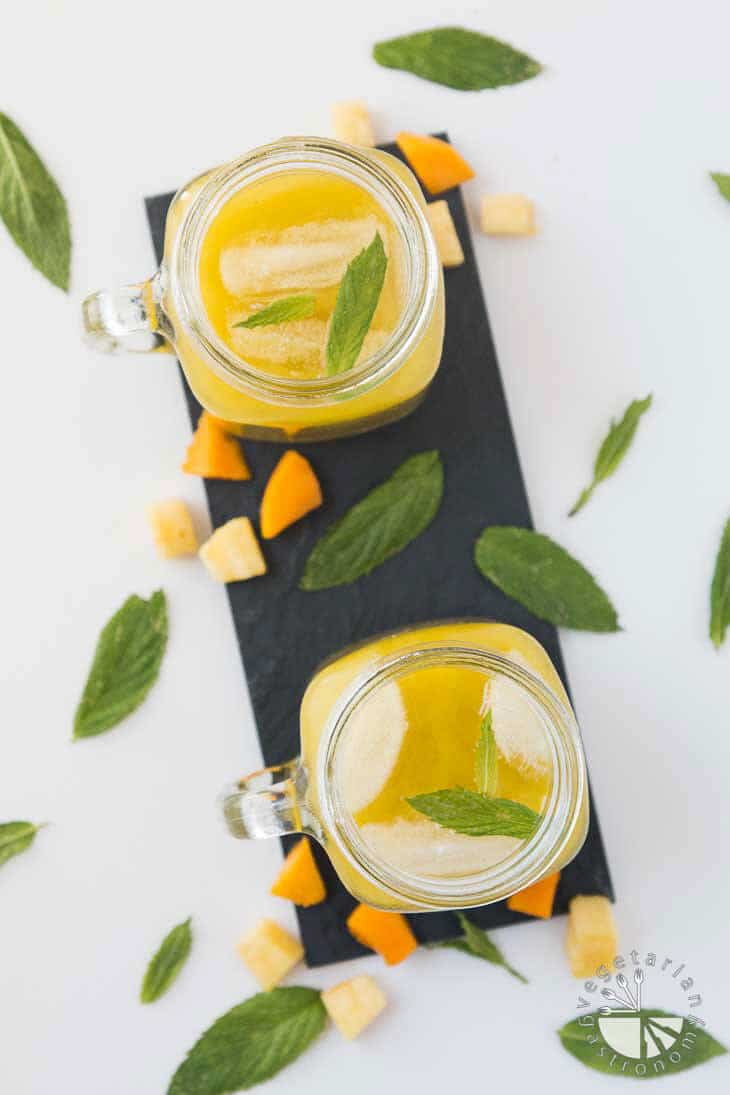 An overhead photograph of two mason jars filled with pineapple mango agua fresca recipe. They are sitting on a rectangular black palate with fruit garnished around the sides.